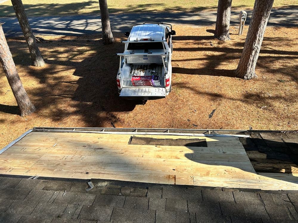 Roof repair process with wooden panels and a parked truck under pine trees.